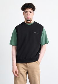 Čierny bezrukávový sveter s výstrihom do V na zelenom tričku, ktorý má malé biele logo na hrudi. Nosené s khaki nohavicami.