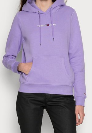 Osoba nosząca lawendową bluzę z kapturem Tommy Jeans z kieszenią z przodu i czarne spodnie, ręka częściowo w kieszeni.