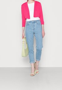 Pink cropped cardigan, white shirt, light blue high-waisted jeans, green handbag, and green strappy heels. Modern casual outfit.
