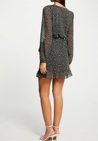 Vestido negro transparente con lunares blancos, mangas largas, dobladillo con volantes y cinturón en la cintura. Combinado con tacones de tiras de color claro.