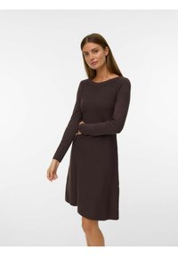Žena s ravnom smešnom kosom koja nosi duge rukave haljinu do kolena tamnosmeđe boje, stoji uz jednostavnu belu pozadinu.