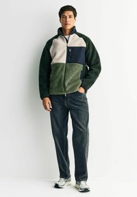 Fleecejakke med grønne, beige og marineblå sektioner; har en lynlåslomme, høj krave og blød tekstur; parret med mørke jeans og sneakers.