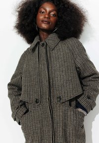 Manteau à motif pied-de-poule en gris et marron, avec un large col, des boutons double boutonnage et des poches latérales. Tissu texturé avec un tissage prononcé.