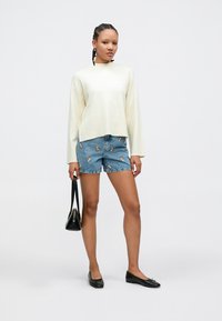 Kremfarget strikkegenser, denimshorts med broderte blomster, svarte flate sko, og liten svart håndveske. Enkel design, ledig passform og lyse farger.