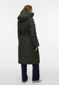 Zaļš pūkains mētelis ar kapuci, jostiņu pie vidukļa, ar ķīļveida rakstu un garu garumu. Valkāts pār tumšu džinsu un gaišām kurpēm.