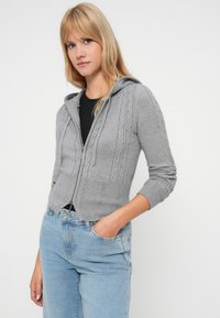 Pelēka kapucīna jumta jaka ar teksturētu kabeli adījumu, ribinātām aprocēm un aukliņu kapuci, kopā ar gaiši zilām džinsu biksēm.