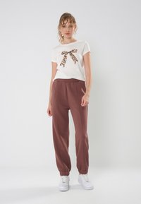 Weißes Baumwoll-T-Shirt mit Leopardenmuster-Bow-Detail, kombiniert mit braunen Jogginghosen mit elastischen Bündchen. Das Modell steht vor einem hellen Hintergrund.