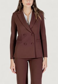 Hnědý dvouřadý blazer vyrobený z hladkého materiálu, s klopami a dvěma předními kapsami, a se šesti odpovídajícími knoflíky. V kombinaci s hnědými kalhotami.