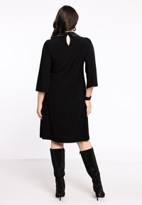 Schwarzes knielanges Kleid mit 3/4-Ärmeln und einem Rückenausschnitt, verziert mit einem dekorativen Kragen mit kontrastierenden Nieten. Kombiniert mit schwarzen kniehohen Stiefeln.