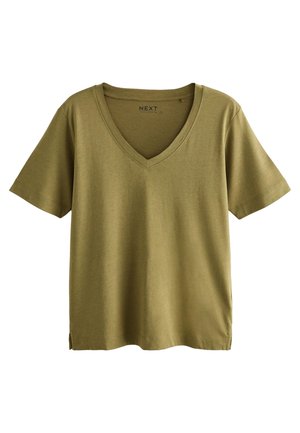 Femme portant un t-shirt vert olive à col en V et un jean ample foncé, une main dans la poche, devant un mur clair uni.