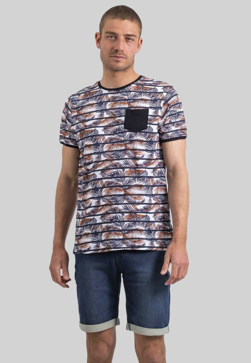 T-shirt à manches courtes avec un motif de feuilles bleues et oranges, fond blanc, col rond et une poche poitrine noire unie. Porté avec un short en denim.