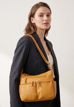 Woman wearing dark blazer over white top carrying tan leather shoulder bag with multiple zippered pockets against plain background.