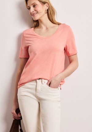 Kobieta w koralowej koszulce z krótkim rękawem i kremowych spodniach, uśmiechająca się, z jedną ręką w kieszeni, a w drugiej trzymająca brązową torebkę.