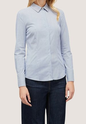 Vrouw draagt een aansluitend blauw-wit gestreept overhemd met knopen, lange mouwen en donkerblauwe spijkerbroek.