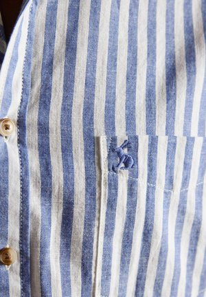 Blau-weiß gestreifter Stoff mit einem kleinen Taschenfach und einem blau bestickten Vogel nahe der oberen rechten Ecke der Tasche.