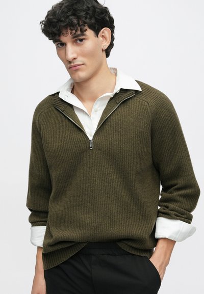 Brauner, gerippter Strickpullover mit einem Reißverschlusskragen über einem weißen Hemd. Die Ärmel sind hochgekrempelt, und der Pullover hat eine legere Passform.