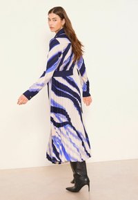 Langes plissiertes Kleid mit diagonalen blauen und cremefarbenen Streifen, maßgeschneidertem Kragen, Knopfdetails und kombiniert mit kniehohen schwarzen Stiefeln.