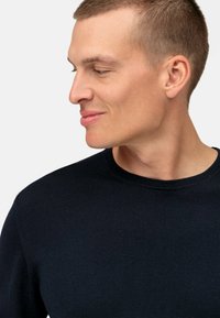 Maglione lavorato a maglia di colore blu navy scuro, con scollatura rotonda, forma aderente e texture a coste, indossato da una persona con capelli corti vista di profilo.