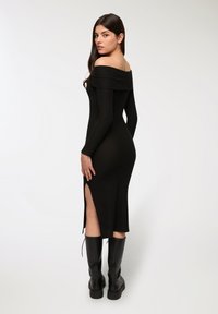 Abito off-the-shoulder nero a coste con maniche lunghe, dotato di uno spacco laterale. Abbinato a stivali neri al ginocchio. Tessuto liscio.