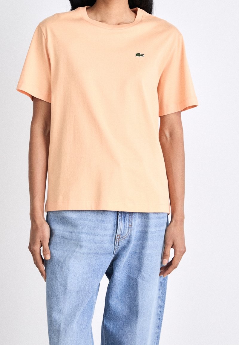 Femme portant un t-shirt uni pêche à manches courtes avec un petit logo crocodile vert et un jean denim bleu clair, debout devant un fond blanc.