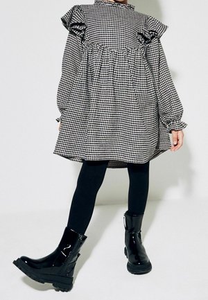 Robe oversize en pied-de-poule noir et blanc avec des manches volantes, assortie à des bottines en cuir verni noir et des collants noirs.