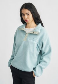 Νεαρή γυναίκα με μακριά σκούρα μαλλιά, φορώντας γαλάζιο φλις πουλόβερ με φερμουάρ στην τσέπη, στέκεται με τα χέρια στις τσέπες μαύρου παντελονιού μπροστά σε λευκό φόντο.