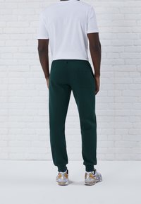 Man i mörkgröna joggingsbyxor, vit kortärmad skjorta och silverfärgade sneakers står vänd mot en vit tegelvägg.