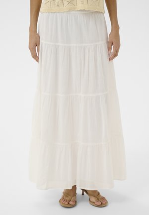 Femme portant une jupe maxi blanche à volants, un haut crocheté beige et des sandales à talons marron clair, debout devant un fond blanc.