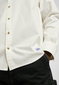 Camisa de color blanco roto con un acabado texturizado, que presenta botones dorados, un dobladillo recto y un parche de logo azul en la esquina inferior izquierda.
