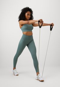 Bustier sportiv verde cu nervuri și colanți înalți, benzi de rezistență elastice cu mânere negre, purtate cu pantofi sport albi.