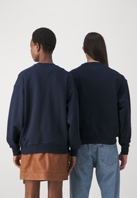 Sudaderas azul marino con puños y dobladillos acanalados, que presentan un corte relajado. Un modelo lleva una falda de cuero marrón; el otro, unos jeans de color azul claro.