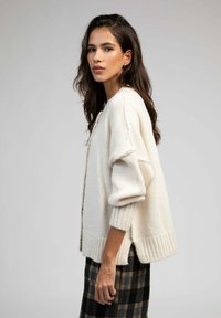 Cardigan en tricot couleur crème avec des poignets et un ourlet côtelés, présentant un devant ouvert et des fentes latérales, associé à une jupe à carreaux.