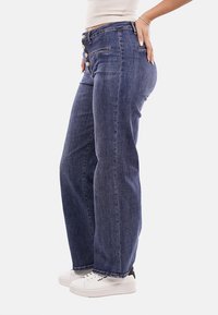 Jeans in denim blu scuro a vita alta con una vestibilità rilassata, caratterizzati da una chiusura con bottoni e due tasche frontali, abbinati a sneakers bianche.
