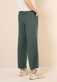 Pantalons verts à jambe large en tissu léger, dotés de poches latérales et d'une coupe décontractée. Associés à des chaussures de sport beiges.