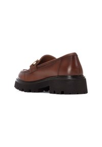 Zapato mocasín de cuero marrón con suela de goma negra gruesa y detalle de hardware dorado en la parte superior. Textura suave con diseño minimalista.