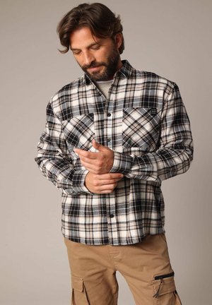 Homme ajustant le poignet d'une chemise à carreaux noire, blanche et beige avec poches avant, portant un pantalon cargo beige sur un fond uni.