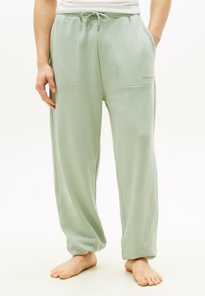 Person trägt hellgrüne Jogginghosen mit Tunnelzug und Fronttaschen, steht barfuß vor einem schlichten weißen Hintergrund.