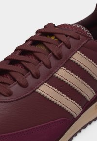 Μπορντό αθλητικό παπούτσι με μπεζ ρίγες με ζιγκζαγκ άκρες, κορδόνια σε ίδιο μπορντό χρώμα και λεπτομερή ραφές πάνω σε επιφάνειες από δέρμα και σουέτ.