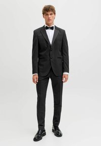 Mladić stoji u crnom smokingu s bijelom košuljom, crnom leptir-mašnom i crnim svečanim cipelama na običnoj bijeloj pozadini.