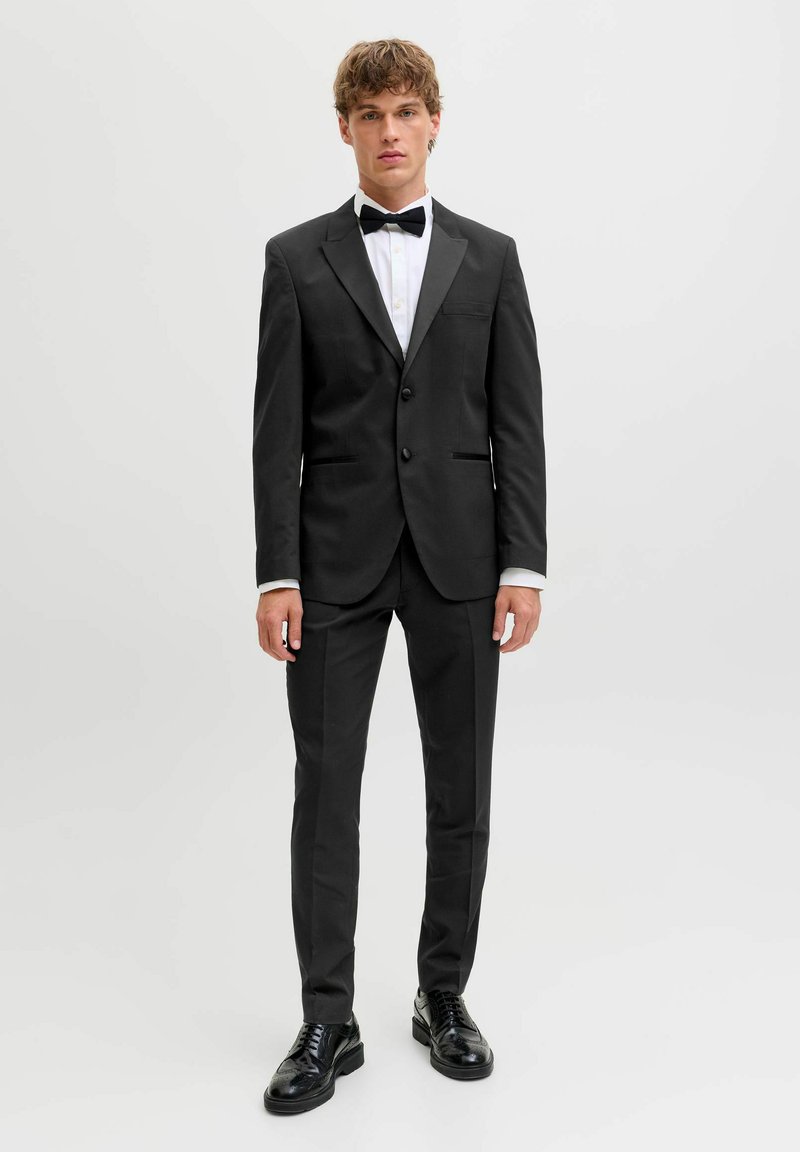 Mladić stoji u crnom smokingu s bijelom košuljom, crnom leptir-mašnom i crnim svečanim cipelama na običnoj bijeloj pozadini.