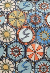 Textil estampado que presenta formas hexagonales en azul, naranja y crema, con motivos de estrellas geométricos y el texto "Gattinoni Desde 1966."