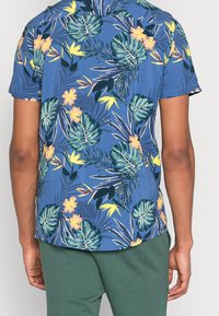Camisetta a maniche corte con sfondo blu e motivi floreali e fogliari tropicali multicolori, realizzata in tessuto leggero.