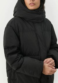 Čierna puffa bunda s vysokou, prehnutou kapucňou; prešívaná textúra; zapínanie na gombíky; elastické manžety; s malou značkovou nálepkou na rukáve.