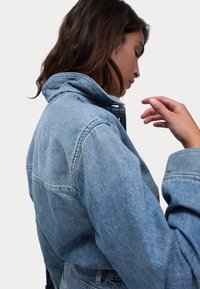 Giacca in denim azzurro chiaro con colletto alto, cuciture prominenti e vestibilità comoda. La texture appare morbida e consumata. Dettagli di cucitura visibili.