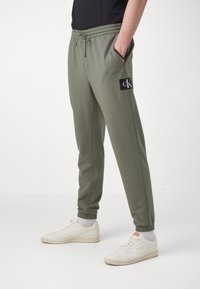 Olivegröna joggerbyxor gjorda av mjukt tyg, med elastisk midja, fållade anklar och en svart logotyp-etikett på vänster lår.