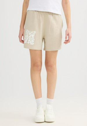 Persona con pantalones cortos beige con texto blanco "living life loving life" en el muslo izquierdo, calcetines blancos, zapatillas blancas y camiseta blanca.