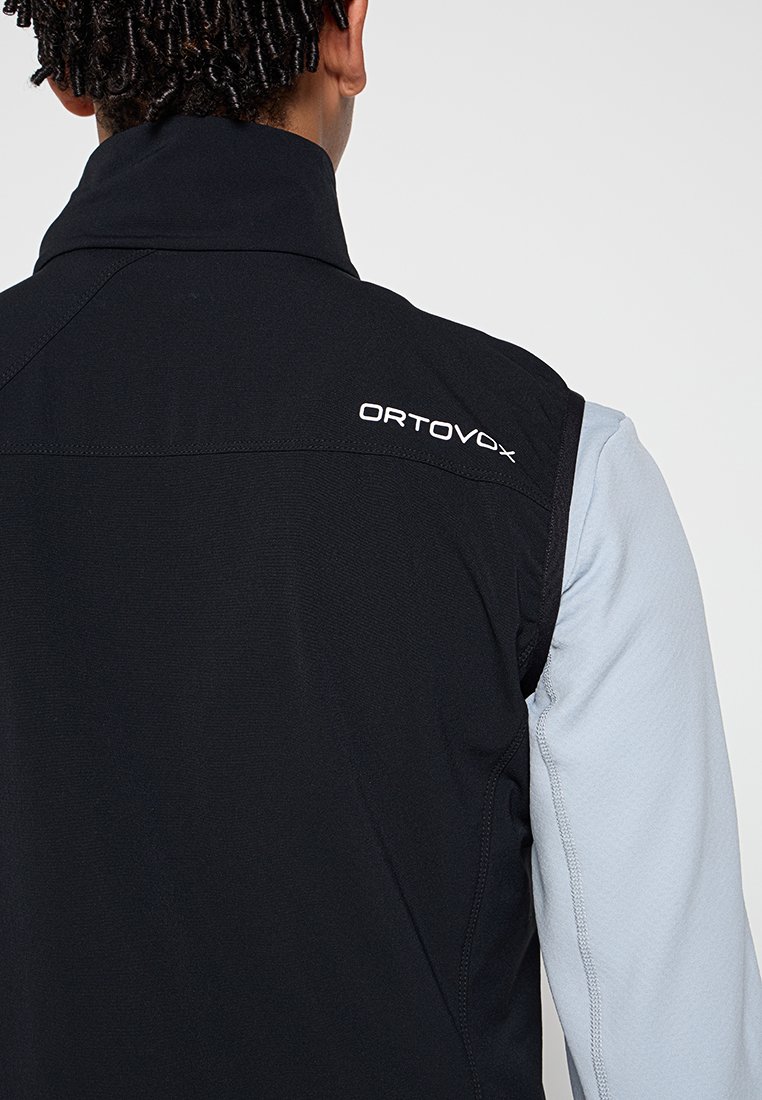 Person, die eine schwarze Ortovox ärmellose Weste über einem hellgrauen Langarmshirt trägt, von hinten gesehen und am Hals beschnitten.
