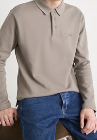 Light brown long-sleeve polo shirt with a textured collar, three golden buttons, and a subtle logo on the chest, paired with blue jeans.