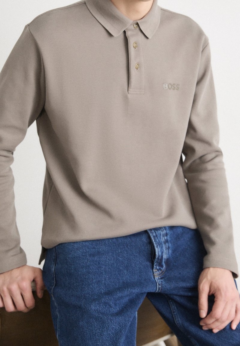 Light brown long-sleeve polo shirt with a textured collar, three golden buttons, and a subtle logo on the chest, paired with blue jeans.