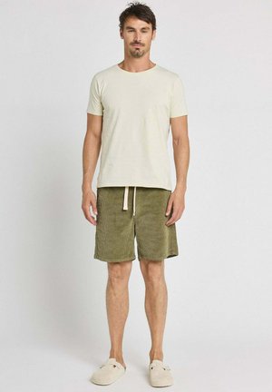 Homme debout face à l'avant portant un t-shirt beige, un short vert olive avec cordon et des chaussures légères beige clair, sur un fond uni.